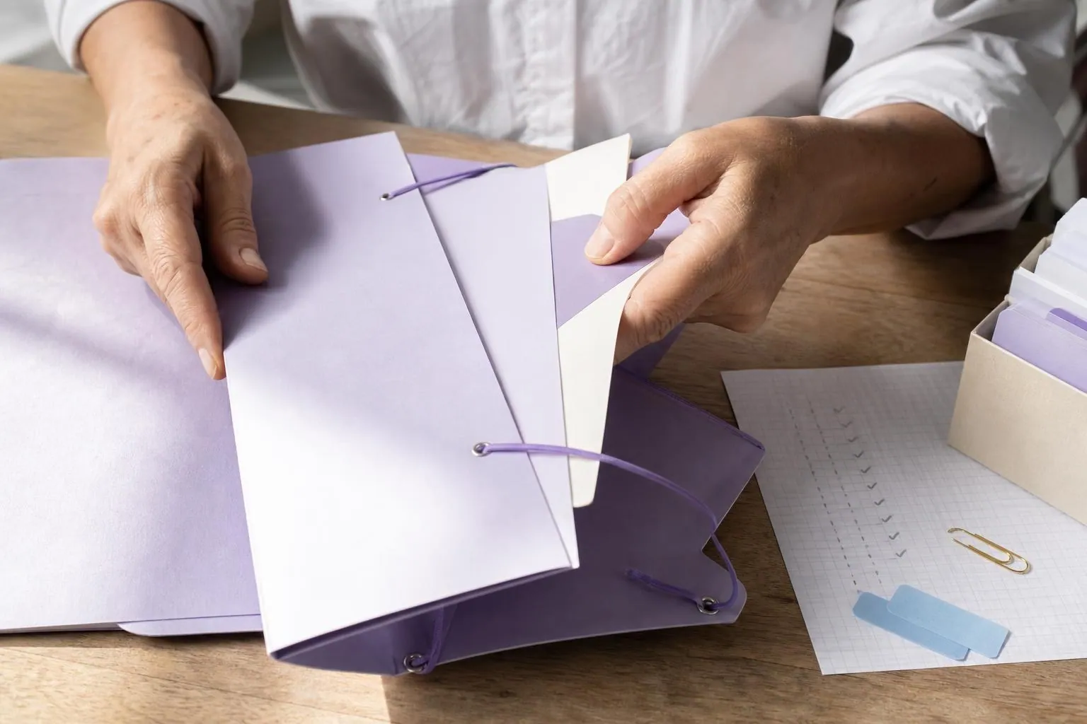 Gros plan vertical sur les mains d’une femme rangeant des chemises cartonnées lilas dans une pochette à élastiques, à côté d’une liste manuscrite cochée